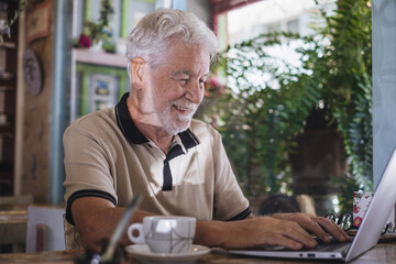 Smiling senior bearded man sitting in cafeteria for a coffee break while using laptop, elderly attractive digital nomadic man typing on keyboard browsing internet