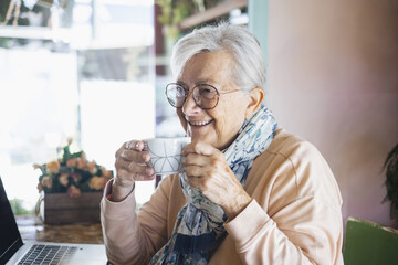 Happy white haired senior woman wearing eyeglasses sitting indoor using laptop while has a coffee break, addicted  elderly lady enjoying a cup of coffee