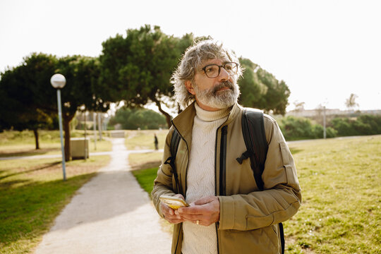 An elderly White man with a white beard and glasses wears a beige jacket and white sweater while standing in a park. He holds a smartphone and looks thoughtful to the right.