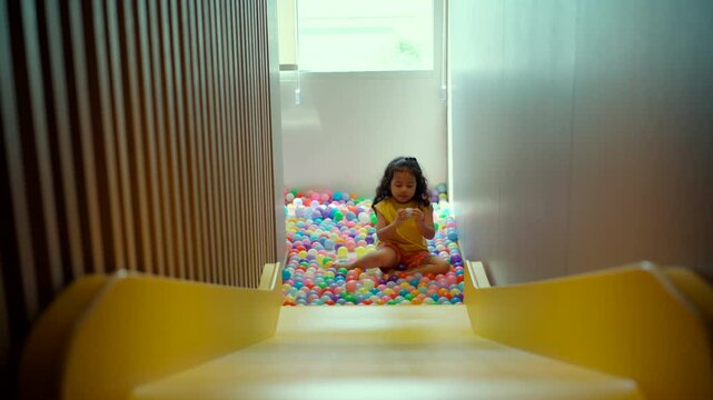 A happy little girl slides down a yellow slide in a kid's club or playroom, enjoying the fun and developing her skills through active play and physical movement.
