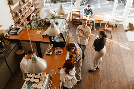 From a high angle inside a sunlit cafe, casually dressed White adult customers and workers stand talking at the wooden counter, while a White man in his 40s sits at a table using his phone nearby.