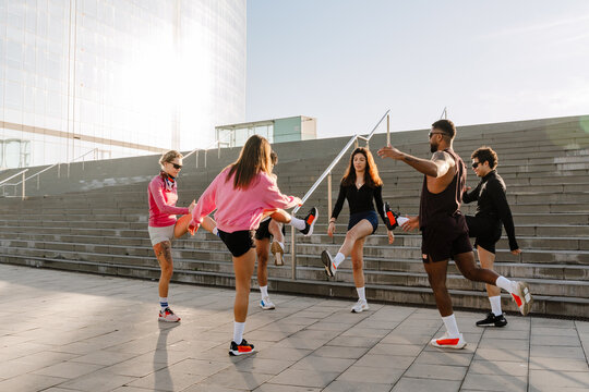 Six multiethnic friends in their late 20s, including men and women wearing athletic clothing doing dynamic leg stretches together in a circle formation on a sunny concrete plaza near large stairs. - Powered by Adobe