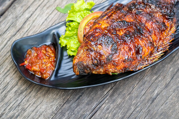 Close-up of grilled fish (ikan bakar) on a black plate with sambal, lettuce, and tomato, set against a rustic wooden table.