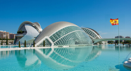 A futuristic landmark in Valencia stands under a blue sky, with the Spanish flag waving.

