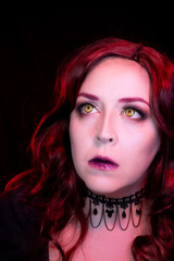 Mysterious woman with gothic vampire makeup and red hair, wearing dark lipstick and a black lace choker. Dramatic lighting highlights her intense eyes and ethereal beauty. 