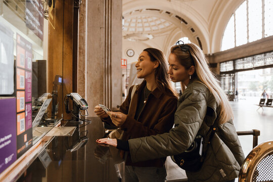 A view from the side of a young Caucasian brunette woman aged 25, buying tickets in a train station with a wide smile on her face, as her mid-20s White, blond female friend stands beside her