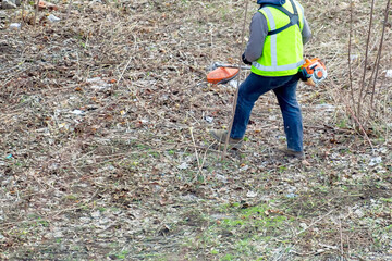 A brush cutter and hedge trimmer in action: a gardener cuts dry stems of grass and bushes on the plot. Working on the ground helps get rid of dry leaves and grass.
