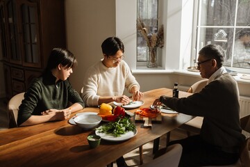Two adult Asian brothers, the older in his late 20s and the younger in his early 20s, cutting veggies together with their elderly grandpa as they sit at home, near a window, in the morning