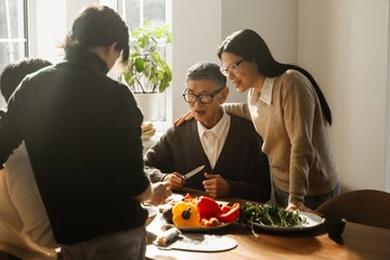 A middle-aged Asian mother in her 50s, standing next to the table where her old father and her two adult sons cut some veggies, and looking at the process, in the morning, at home