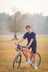Fototapeta premium Handsome young man in a blue shirt and black sports shorts is happily with a red bicycle in the morning for exercise, standing and posing on a green lawn in the park. Healthy exercise.