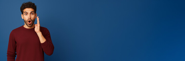 Surprised young man in a burgundy sweater holding his hand to his face against a blue background.