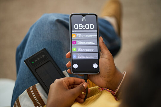 Close-up view of woman holding smartphone showing health reminders on screen. Device connected to solar charger, human hand interacting with screen, wearing casual clothing
