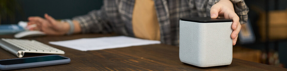 Boy adjusting smart speaker on wooden desk, with blurred background showing keyboard and phone....