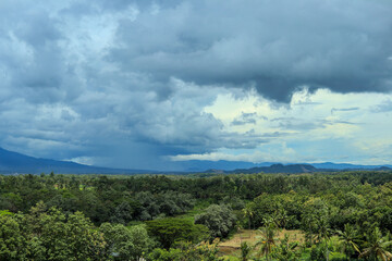 A wide-angle shot of a lush green forest valley stretching to a distant mountain range, with a dramatic sky of dark, stormy clouds overhead.