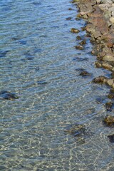 Transparent Coastal Waters and Rocky Shoreline with Sunlight Reflections; crystal-clear coastal water reveals submerged rocks and seaweed near a rugged stone shoreline