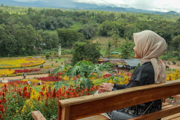A woman wearing a beige hijab sits on a wooden bench, gazing out at a colorful flower garden.