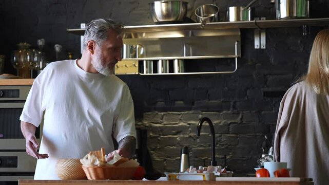 A White elder blonde woman is giving a glass of water to her husband in the kitchen, then she takes a bowl and brings it to the stove while he places the glass down and talks to her.