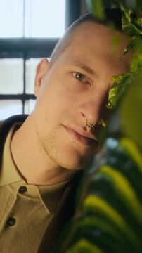 Vertical closeup portrait of young Caucasian man with nose piercing leaning his face against green wall with leaves and posing for camera with deep gaze