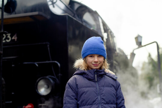 Child in blue beanie stands smiling by a steaming black locomotive outdoors.
