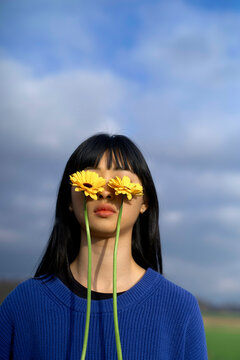 Woman with long hair holds two yellow flowers against her eyes under a cloudy sky. Brandenburg, Germany