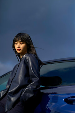 Woman in leather jacket leans against a blue car under a cloudy sky. Brandenburg, Germany