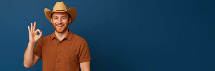 Smiling man in a cowboy hat and brown shirt making an OK hand gesture against a blue background