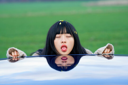 Woman playfully sticking her tongue out at her reflection on a shiny car roof. Brandenburg, Germany