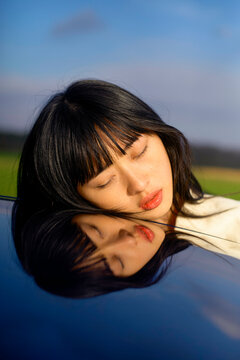 Woman resting her head on a car hood, reflecting in the polished surface against a blue sky. Brandenburg, Germany
