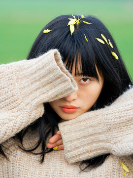 Woman with black hair partially covers face with sweater, yellow petals in hair, green background. Brandenburg, Germany