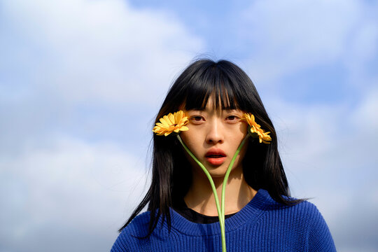 Woman with straight black hair holding two yellow flowers against a blue sky backdrop. Brandenburg, Germany