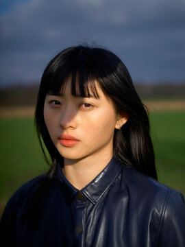 A woman with long black hair wearing a dark leather jacket stands outdoors under a cloudy sky. Brandenburg, Germany
