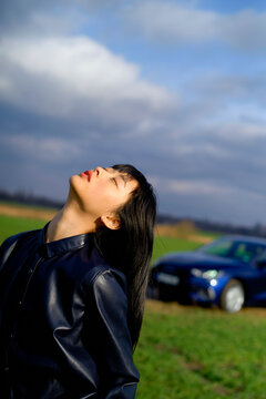Woman in black jacket standing in a field, with a blue car in the background under a cloudy sky. Brandenburg, Germany