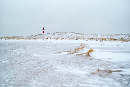 Snow-covered beach with a distant red lighthouse under a grey sky. Sylt, Germany