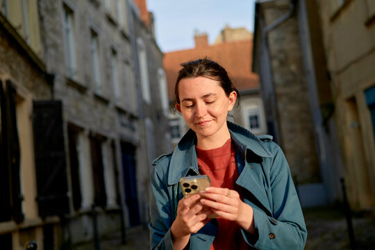 Woman standing on a narrow cobblestone street using a smartphone on a sunny day. Normandy, France