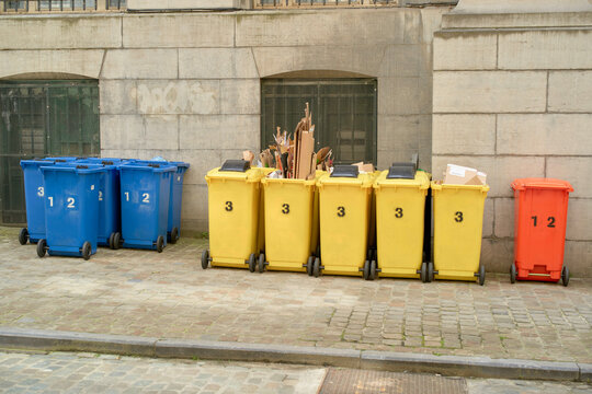 Blue, yellow, and red recycling bins lined up against a stone wall on a cobblestone street. Brussels, Belgium
