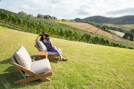 Woman in hat relaxing on chairs in vineyard with rolling hills in the background Waiheke Island, Auckland, New Zealand