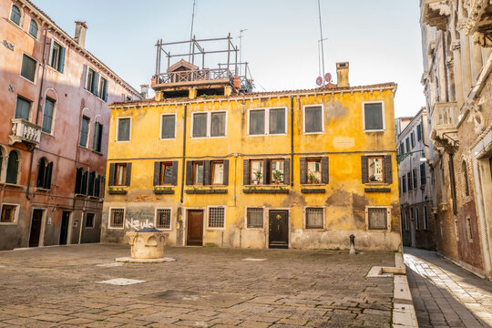 Yellow building with weathered facade, small square, and fountain under clear blue sky. Venice, Italy