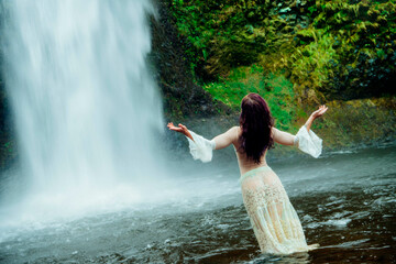 Woman standing in water near a waterfall, arms raised, surrounded by lush greenery. Silver Falls, Oregon, USA