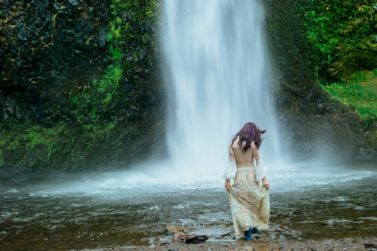 Woman in a flowing dress stands before a tall, majestic waterfall surrounded by lush greenery. Silver Falls, Oregon, USA