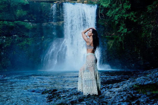 Woman in a bohemian skirt poses by a majestic waterfall in a lush green forest. Silver Falls, Oregon, USA