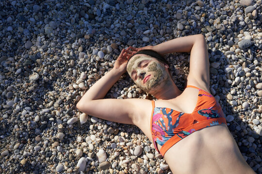 Woman in colorful bikini with mud face mask sunbathing on pebble beach.