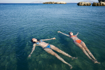 Two women floating on calm sea water under a clear blue sky near rocky shore.