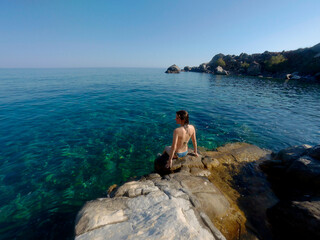 Woman in swimsuit sitting on rocky shore, gazing at clear blue sea under a bright sky.