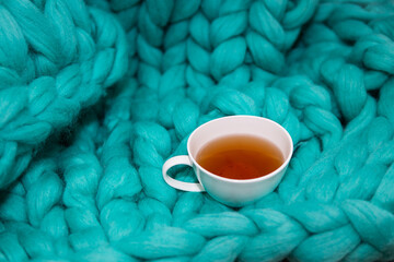 The concept of coziness and comfort is a green knitted blanket on which there is a white cup of tea. Close-up.