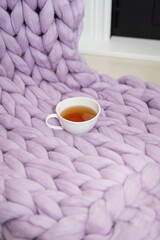 The concept of coziness and comfort is a pink-lavender knitted blanket with a white cup of hot tea on it. Place for inscription, vertical photo.