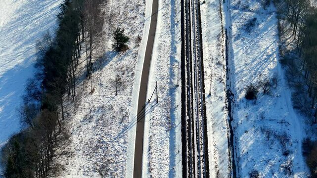 An aerial top-down view of railway tracks and a parallel road slicing through a snow-covered forest. Bare trees cast long shadows over the frozen landscape in winter light.