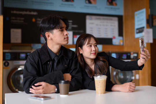Young Asian couple sitting in laundry shop drinking coffee and using phone