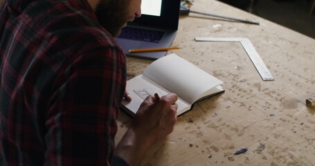 handsome serious man of European appearance with beard, in plaid shirt, makes a pencil drawing in a notebook with a ruler, in a carpentry workshop, video filming from behind