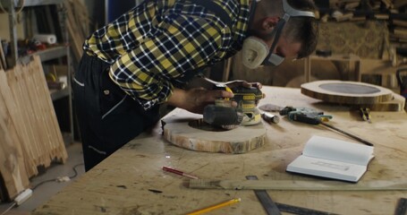 a man of European appearance in protective headphones and a mask processes a wooden saw cut using a tool, sanding wood with a flex, work in a carpentry workshop