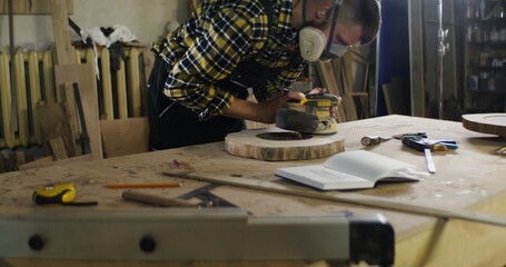 a man of European appearance in protective headphones and a mask processes a wooden saw cut using a tool, sanding wood with a flex, work in a carpentry workshop